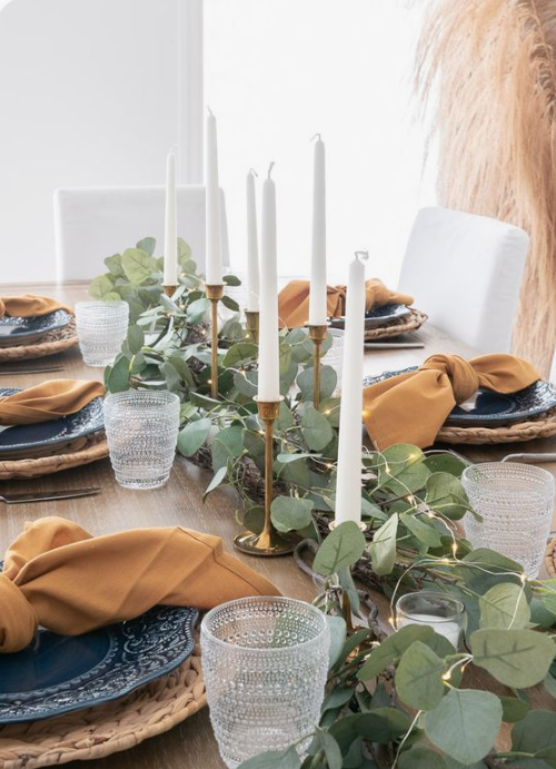 a very simple fall tablescape with a greenery and lights runner, tall candles, woven placemats, blue plates and rust napkins