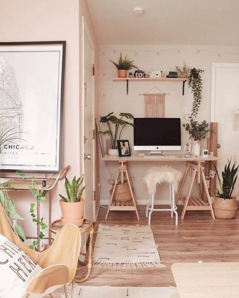 A warm colored boho space with a wooden trestle desk, an open shelf, potted greenery and plants and long fringe