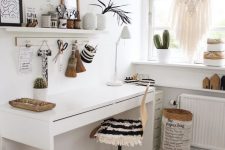 a white boho home office with open shelves, a white desk, a chair with a fringe cover, a boho ottoman, a macrame hanging and potted plants