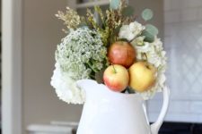 an arrangement of a white jug, white blooms, greenery, apples and a copper mug for simple and chic fall decor