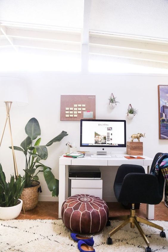 an attic boho chic home office with a white desk, a leather ottoman, a black chair, potted plants on the floor and some on the walls