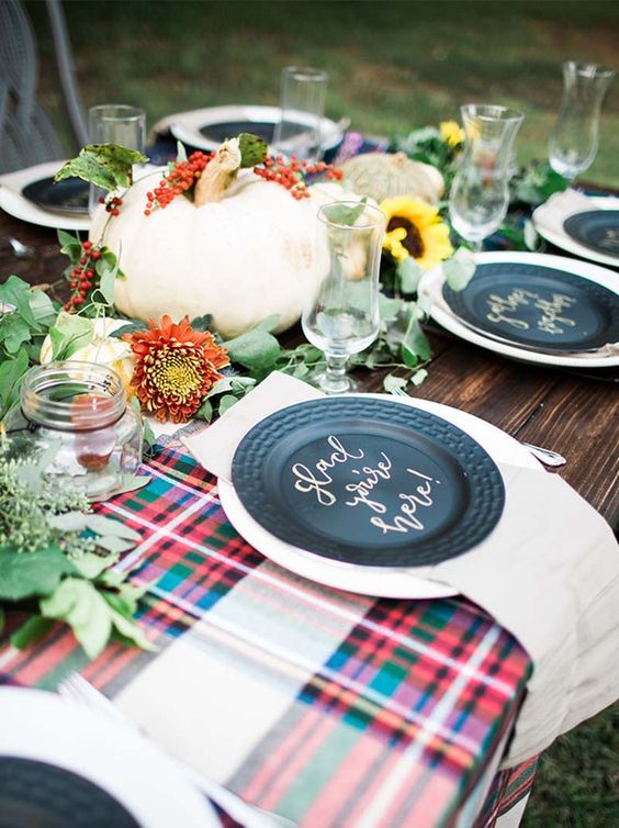 a bright fall table setting with plaid fabric, blakc plates, blooms, greenery and pumpkins looks very cute
