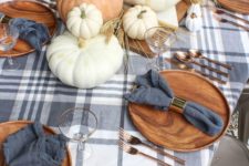 a cozy rustic fall tablescape with large pumpkins stacked, wheat, slate grey napkins and a plaid tablecloth, copper cutlery and wooden chargers