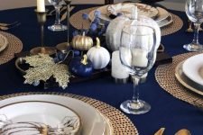 a bold Thanksgiving table with gold cutlery, woven chargers, printed plates, blue, gold and white pumpkins and candles