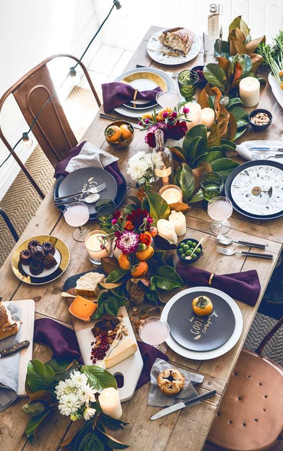 a bright Thanksgiving table setting with an uncovered table, a greenery and fruit runner, candles, blooms and black chargers