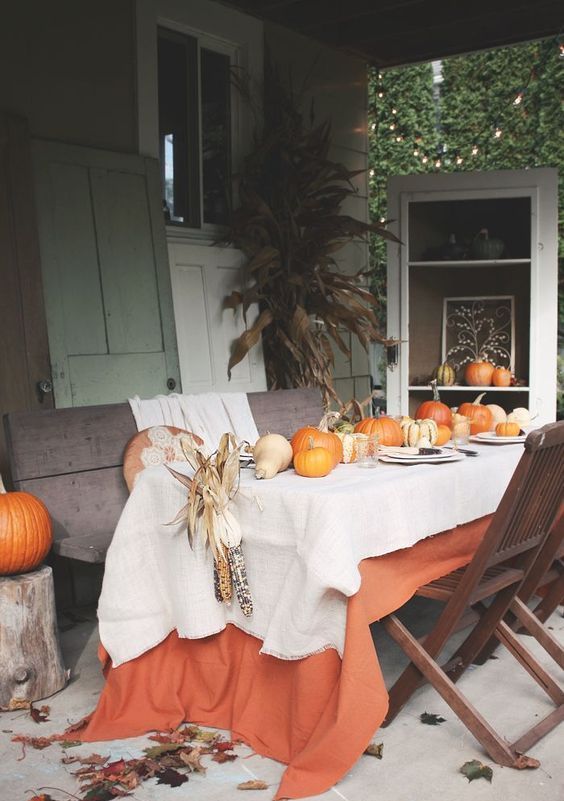 a bright fall tablescape with a white and an orange tablecloth, pumpkins, corn cobs and plates and placemats