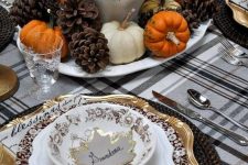 a cozy rustic vintage Thanksgiving tablescape with a plaid tablecloth, quote napkins, woven chargers, gold and pritned plates, a chic centerpiece of wheat, pumpkins and pinecones