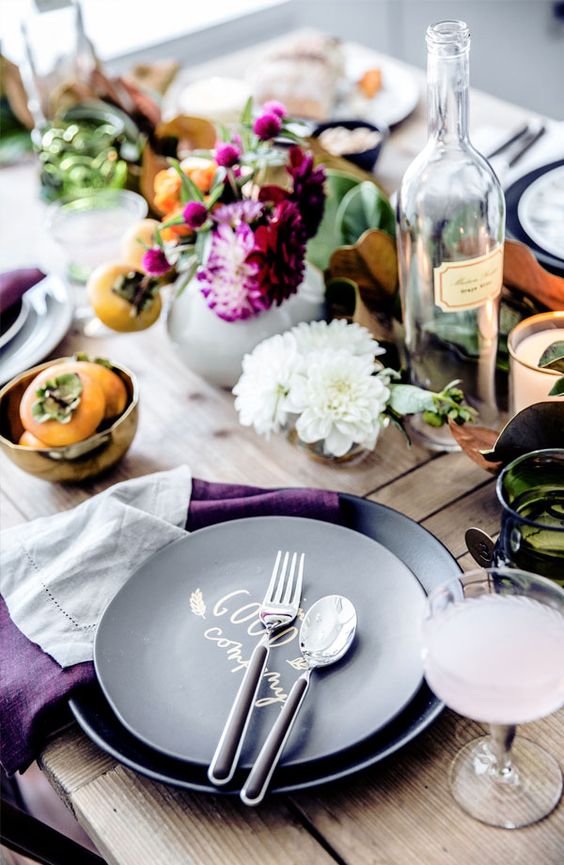 A laid back and rustic Thanksgiving tablescape with greenery, bold purple blooms, fruits, black plates and purple napkins