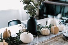 a modern Thanksgiving tablescape with a neutral fabric runner, neutral real and plywood pumpkins, greenery, black plates and striped napkins