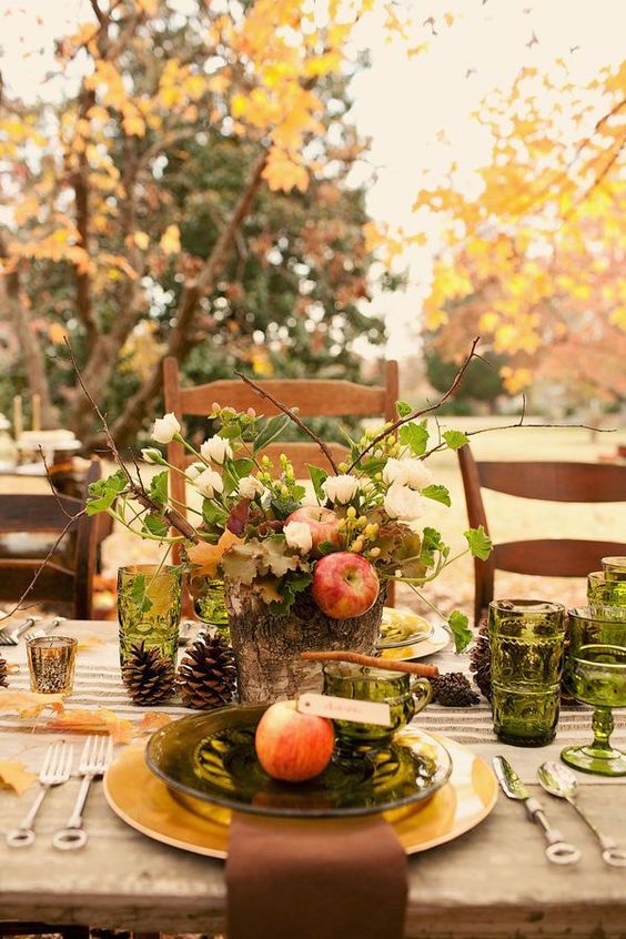 a pretty Thanksgiving table with grene glasses and plates, pinecones, a floral and apple centerpiece in a tree stump