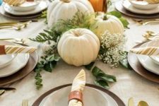 a rustic Thanksgiving table with wooden chargers, a burlap tablecloth, gold cutlery, white pumpkins and greenery plus candles