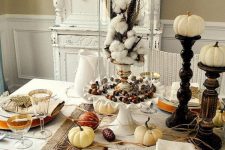 a rustic vintage Thanksgiving tablescape with neutral textiles, a striped runner, white porcelain and wooden stands with pumpkins plus cotton in a cloche
