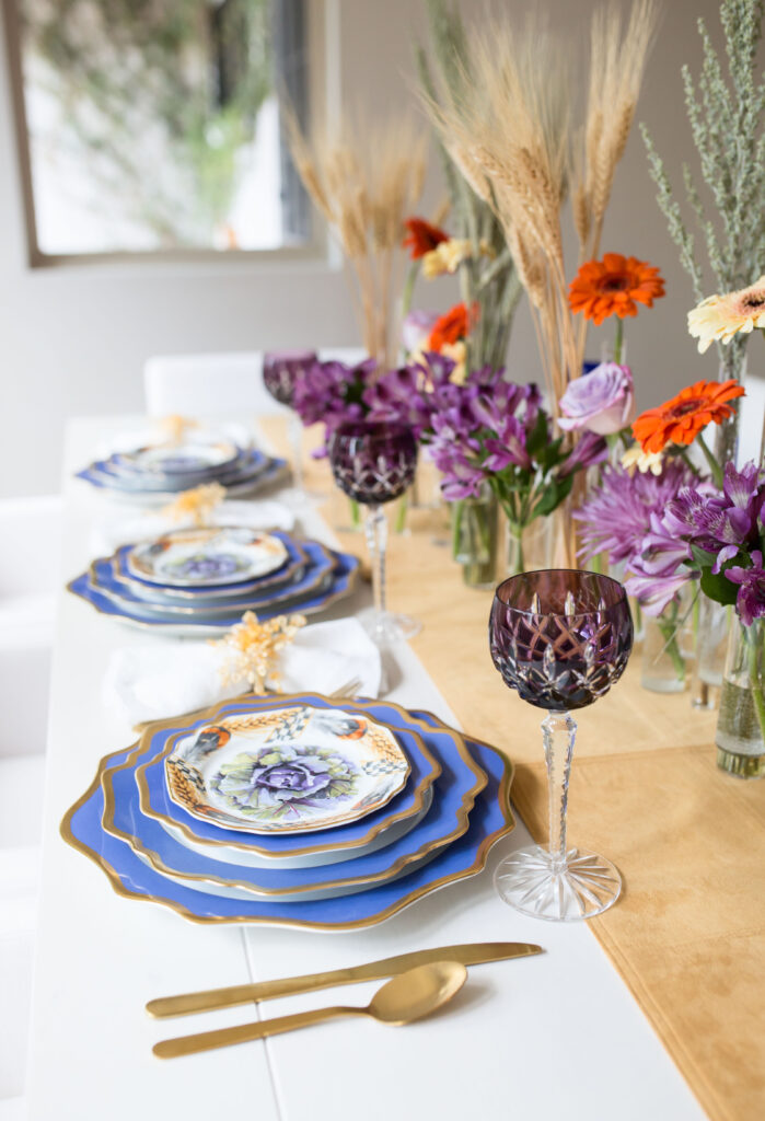 a stylish Thanksgiving table setting with a neutral runner, purple and orange blooms, wheat, blue plates with a gold edge, gold cutlery