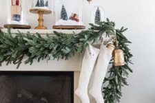 a flocked greenery garland with white stockings and a vintage bell hanging for lovely and chic Christmassy decor