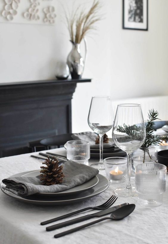 a minimalist Christmas table with grey napkins, evergreens and candles in glass candleholders plus black cutlery for a stylish look