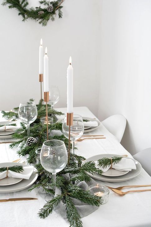 a minimalist and natural Christmas table with an evergrene garland and pinecones, gold cutlery and copper candleholders