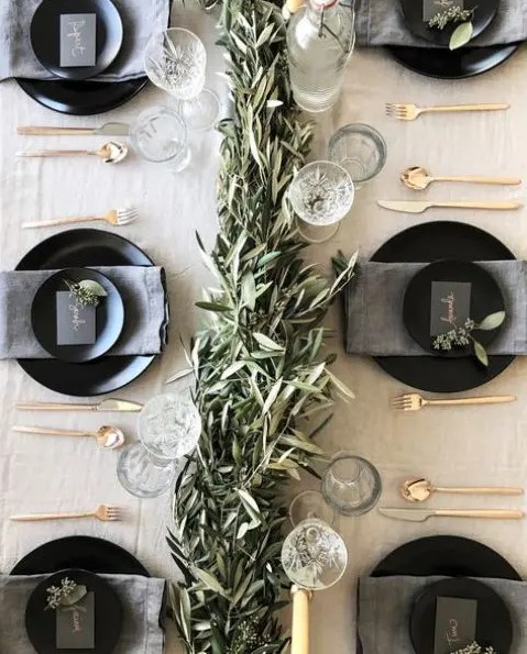 a neutral tablescape with a greenery garland, glasses, matte black plates and chargers and gold cutlery