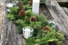 a rustic Christmas tablescape with an uncovered table, an evergreen runner with candles and pinecones is a cozy idea