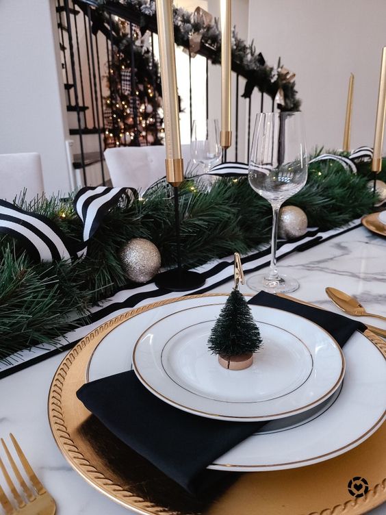 a stylish black and white NYE party table with gold chargers and white plates, black napkins, an evergreen runner with a ribbon and tall and thin candles