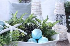 a white bowl with greenery, starfish, pinecones and blue ribbon Christmas ornaments is a lovely Christmas centerpiece for a beach space