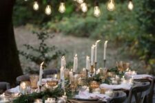an elegant Christmas tablescape with an evergreen runner, tall and thin candles, metallic ornaments and some lights over the table