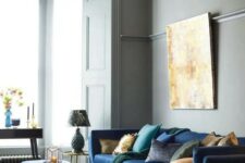 a beautiful and refined living room with grey walls, a navy mid-century modern sectional, a navy pouf and yellow and blue stools, a crystal chandelier and a statement artwork