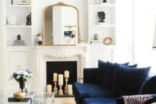 a beautiful neutral living room with a candle fireplace, a modern navy sofa, built-in shelves, a glass coffee table and lovely gold touches here and there