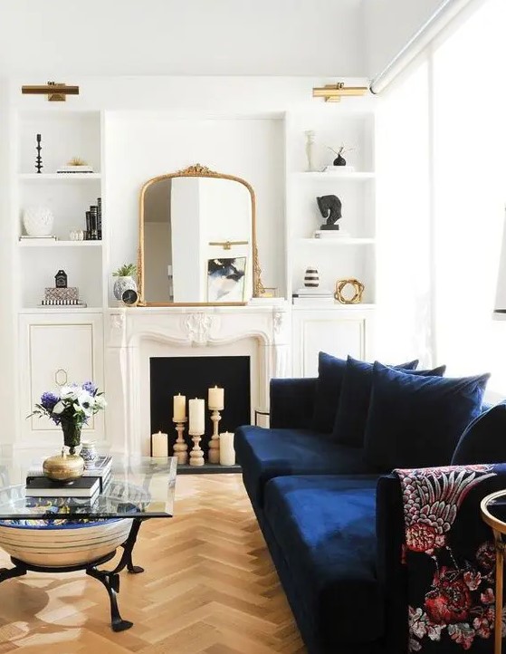 A beautiful neutral living room with a candle fireplace, a modern navy sofa, built in shelves, a glass coffee table and lovely gold touches here and there