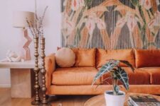 a boho living room with a floral tapestry, an orange sofa, a round table and a side table plus candle holders and a boho rug