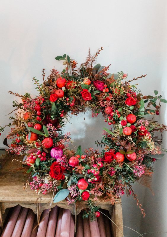 a bold and chic Christmas wreath with greenery and dark foliage, red apples and roses, some moss looks very wild and fresh