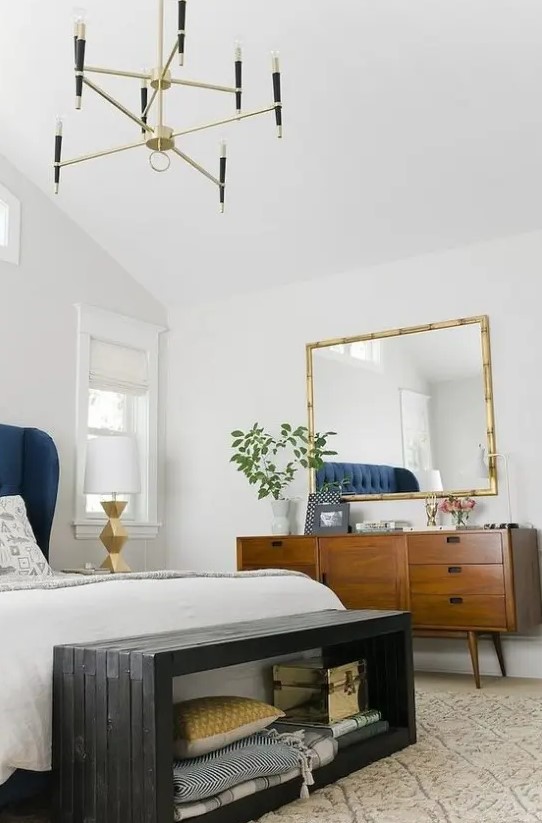 A chic and bright mid century modern bedroom with white walls, rich stained furniture, a black storage bench and a gilded chandelier