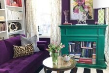 a colorful living room with a depe purple accent wall and a matching sofa, a striped rug, a floral pendant lamp and an emerald fireplace with books
