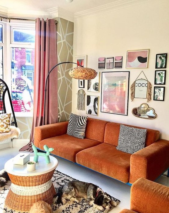 A gorgeous eclectic living room with a modern rust colored sofa and a matching chair, a round table, a pretty gallery wall and a floor lamp