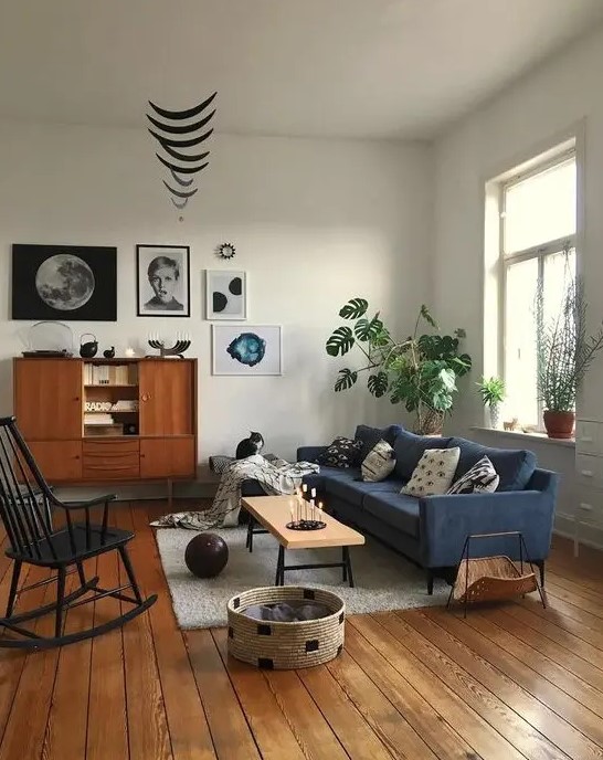 A mid century modern living room with a blue sofa, a stained buffet, a black rocker, a low bench, a basket for storage and a cool gallery wall