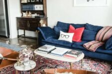a mid-century modern living room with a vintage bureau, a modern blue sofa with colorful pillows, leather chairs, coffee tables and a red printed rug