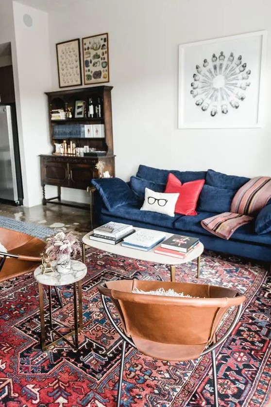 A mid century modern living room with a vintage bureau, a modern blue sofa with colorful pillows, leather chairs, coffee tables and a red printed rug