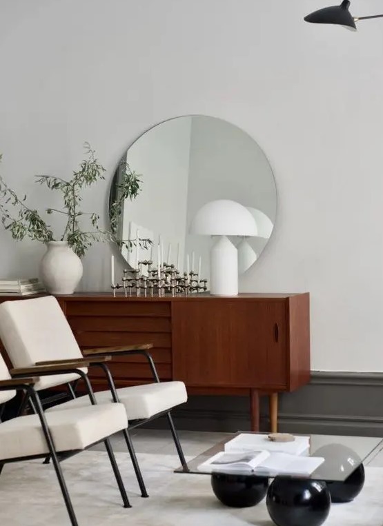 a mid-century modern space with an elegant stained credenza, white chairs, a round mirror, greenery and a coffee table
