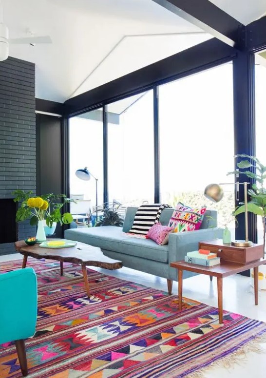 A mid century modern to boho living room with a black brick fireplace, a blue sofa, a turquoise chair, bright pillows and rugs, some potted plants