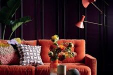 a moody living room with purple paneled walls, a modern orange sofa, orange floor lamps, a gold hammered table and statement plants