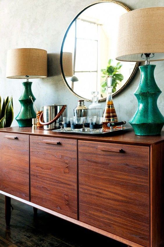 a rich-stained modern credenza with table lamps with emerald bases, as a mini home bar and a round mirror over it