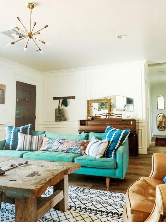 An eclectic living room with a mid century modern turquoise sofa, a rough wooden table, a chair, a wooden dresser and some mirrors on it