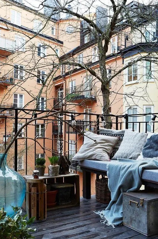 a Nordic balcony with an upholstered bench, crates and baskets, a metal chest fro storing blankets and pillows, potted greenery and lights