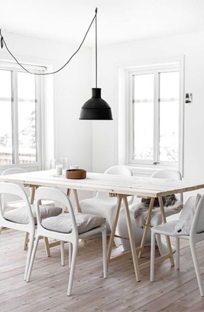 a Nordic dining space whith a whitewashed trestle dining table, matching white chairs, a black pendant lamp and natural light