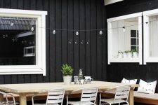 a Scandinavian terrace with a wooden deck, a stained corner bench, a stained wooden dining table and white metal chairs, lights over the table