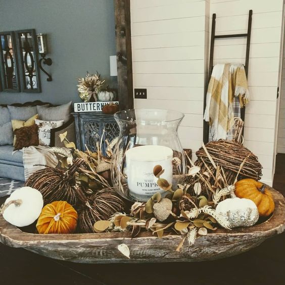 a fall decoration of a weathered dough bowl, fake pumpkins of various materials, dried leaves and a pillar candle