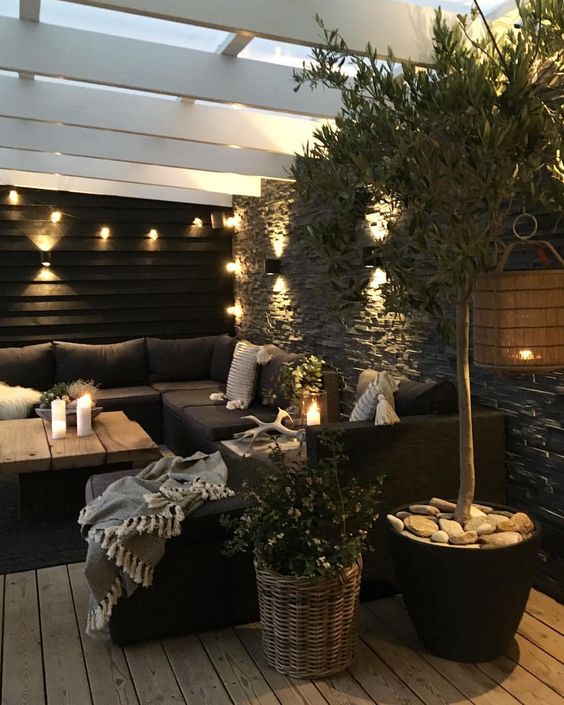 a moody Nordic terrace with a black sectional, a living edge coffee table, potted greenery and candle lanterns and string lights