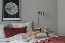 a serene Scandinavian bedroom with grey walls, a bed with grey and deep red bedding, a moon print and brass sconces