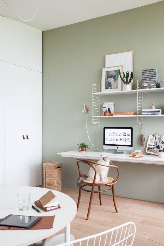 A welcoming Nordic home office with a sage green accent wall, a white wall mounted desk and a shelving unit, a wooden chair and a round table