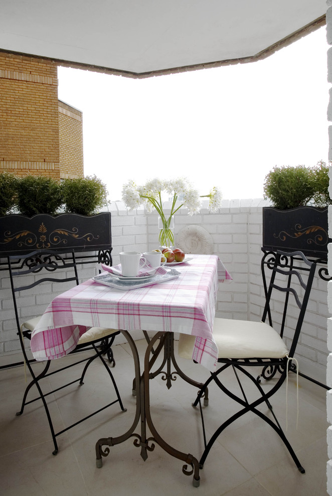 A small vintage style balcony with white brick, a vintage forged table and chairs, potted greenery, a pink plaid tablecloth and some blooms