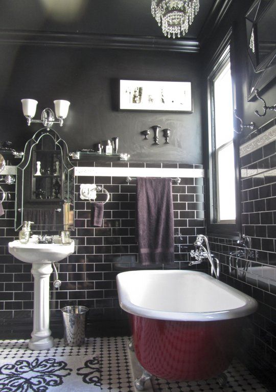 A Gothic bathroom with black walls and glossy black tiles, a fuchsia colored tub, a free standing sink, a chic mirror and a crystal chandelier